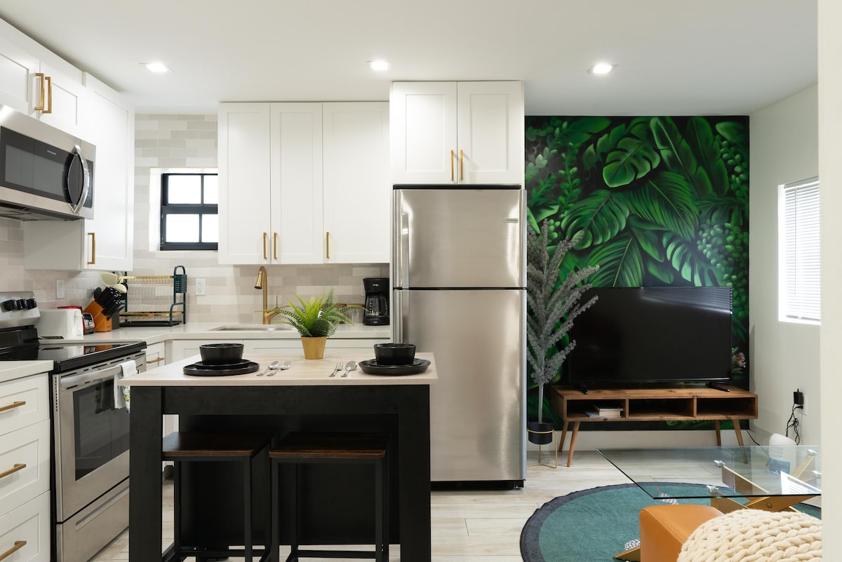 A modern kitchen features stainless steel appliances, including a refrigerator and microwave. The white cabinetry provides ample storage, complemented by a tropical mural on one wall. A dining area with two stools sits adjacent to a coffee maker, while a sleek television is positioned on a wooden stand.