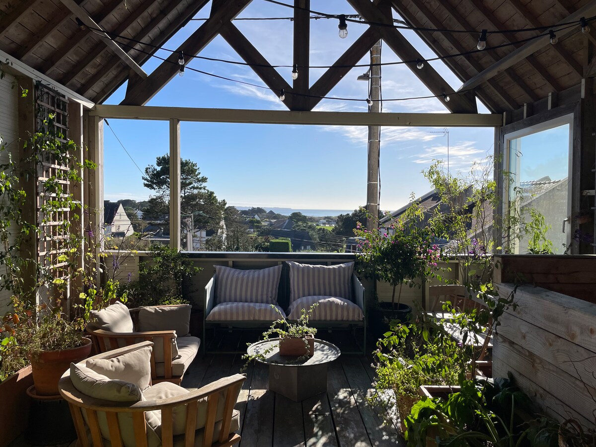 A sunlit terrace is displayed, featuring comfortable seating and various potted plants. Large windows frame a view of the ocean and surrounding greenery, while overhead string lights add a soft ambiance. The wooden structure and natural decor contribute to a relaxing atmosphere.