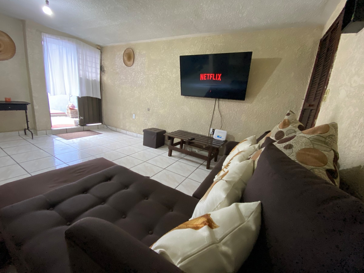 A comfortable seating area is featured with a dark-colored sofa adorned with decorative pillows. A flat-screen TV displaying Netflix is mounted on the wall. Natural light enters through a nearby window, illuminating the tiled flooring.