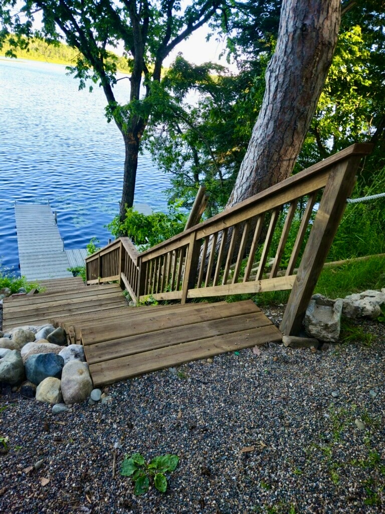A wooden staircase leads down towards the lake, framed by lush greenery. Smooth stones line the pathway, and a wooden dock is visible across the water, creating a direct connection to the serene lakeside environment.