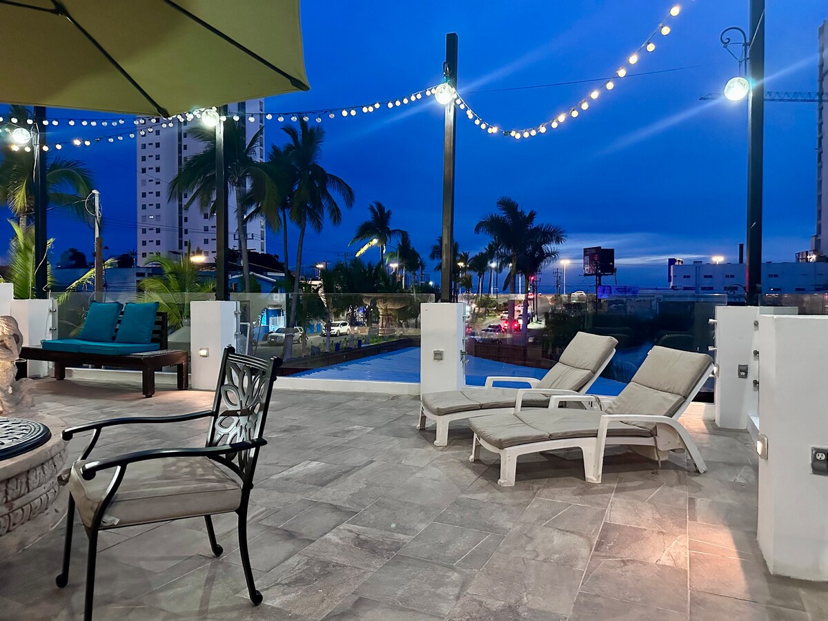 An outdoor terrace is illuminated by string lights against a twilight sky. Lounge chairs are positioned around a seating area, featuring a charcoal BBQ and views of the pool and surrounding palms. The space is inviting, perfect for relaxation and enjoying the evening.