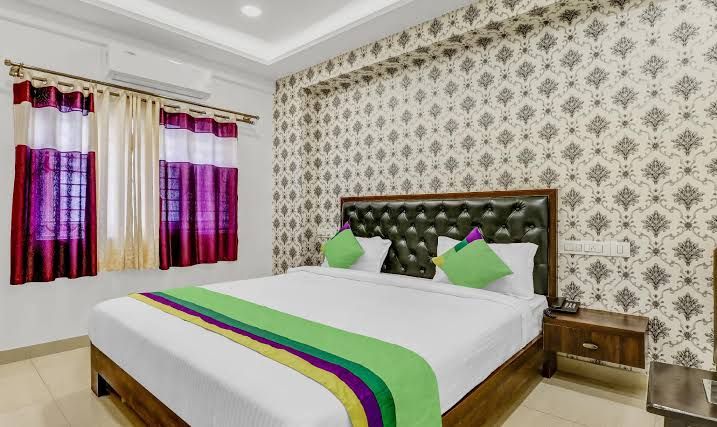 A spacious sleeping area features a large bed with a tufted headboard, accented by bright green pillows. Decorative wallpaper adds a patterned backdrop. Natural light enters through a window with sheer curtains, complemented by red and purple drapes.