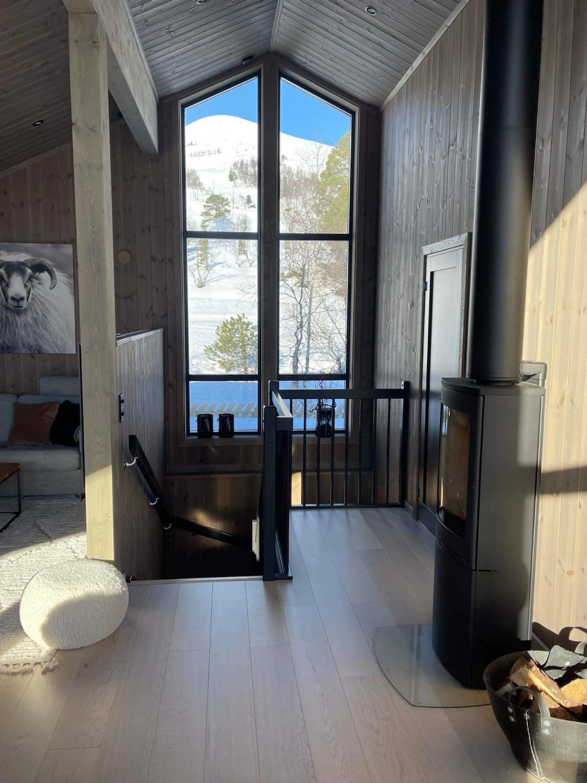 A well-lit living space features tall windows that provide a view of the snowy landscape outside. A modern wood stove is positioned near the stairs, complementing the cozy atmosphere. Natural wood walls enhance the warmth of the interior, while soft textures are present in the furnishings.