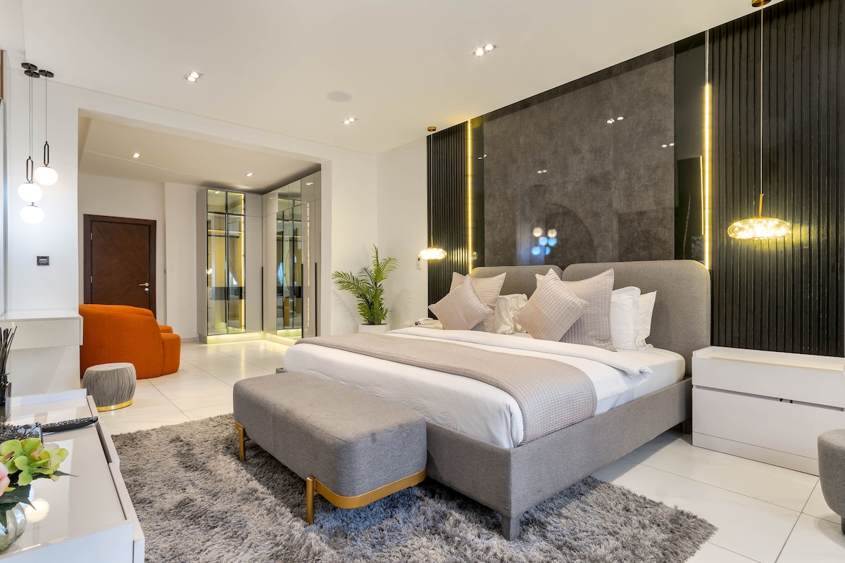 The bedroom features a spacious bed with soft linens and decorative pillows, complemented by a plush area rug. A contemporary design is highlighted by wall-mounted lights and sleek furniture. A mirrored wardrobe is visible, reflecting the room's elegant atmosphere.
