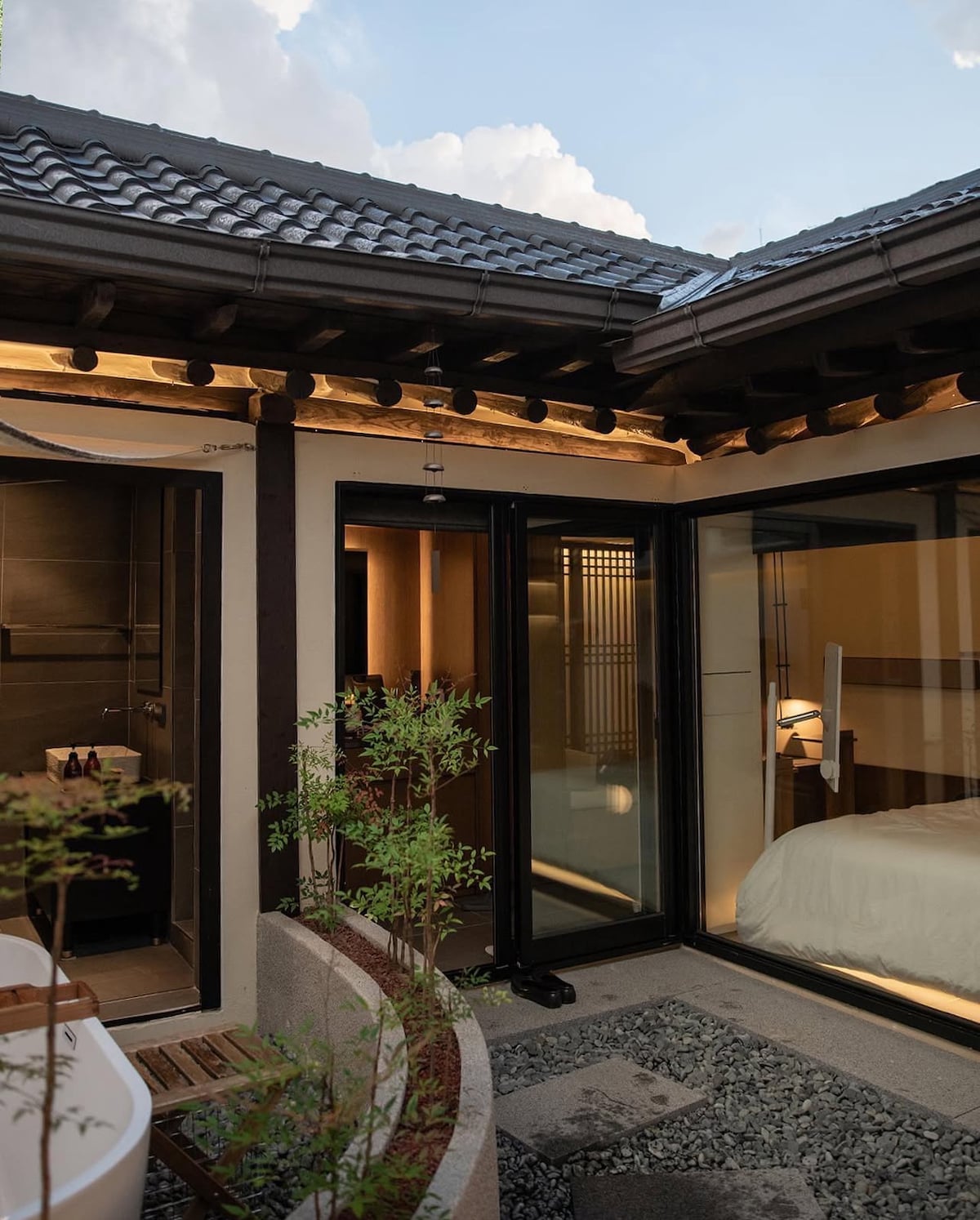 An outdoor area framed by modern glass doors, revealing a harmonious blend of traditional and contemporary architecture. Soft light highlights the spacious bed inside, while a private hot tub is visible nearby, surrounded by natural greenery and pebbles.