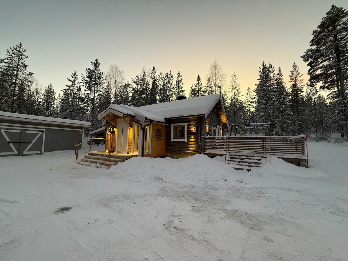 The exterior of the timber cottage is highlighted by fresh snow covering the ground and steps. Soft lights illuminate the entrance, casting a warm glow against the wooden facade. Surrounding pine trees frame the cozy retreat, with a serene winter sky in the background.