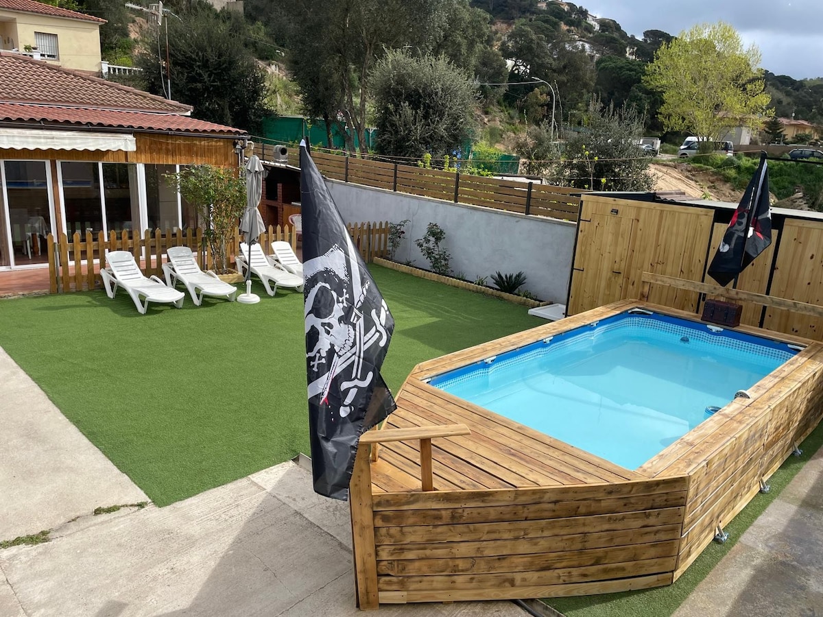 An outdoor space features a private rectangular pool surrounded by a wooden deck. Sun loungers are positioned on the artificial grass area. Flags with a skull motif are visible, and a patio area with a roof can be seen in the background, creating a relaxed setting.