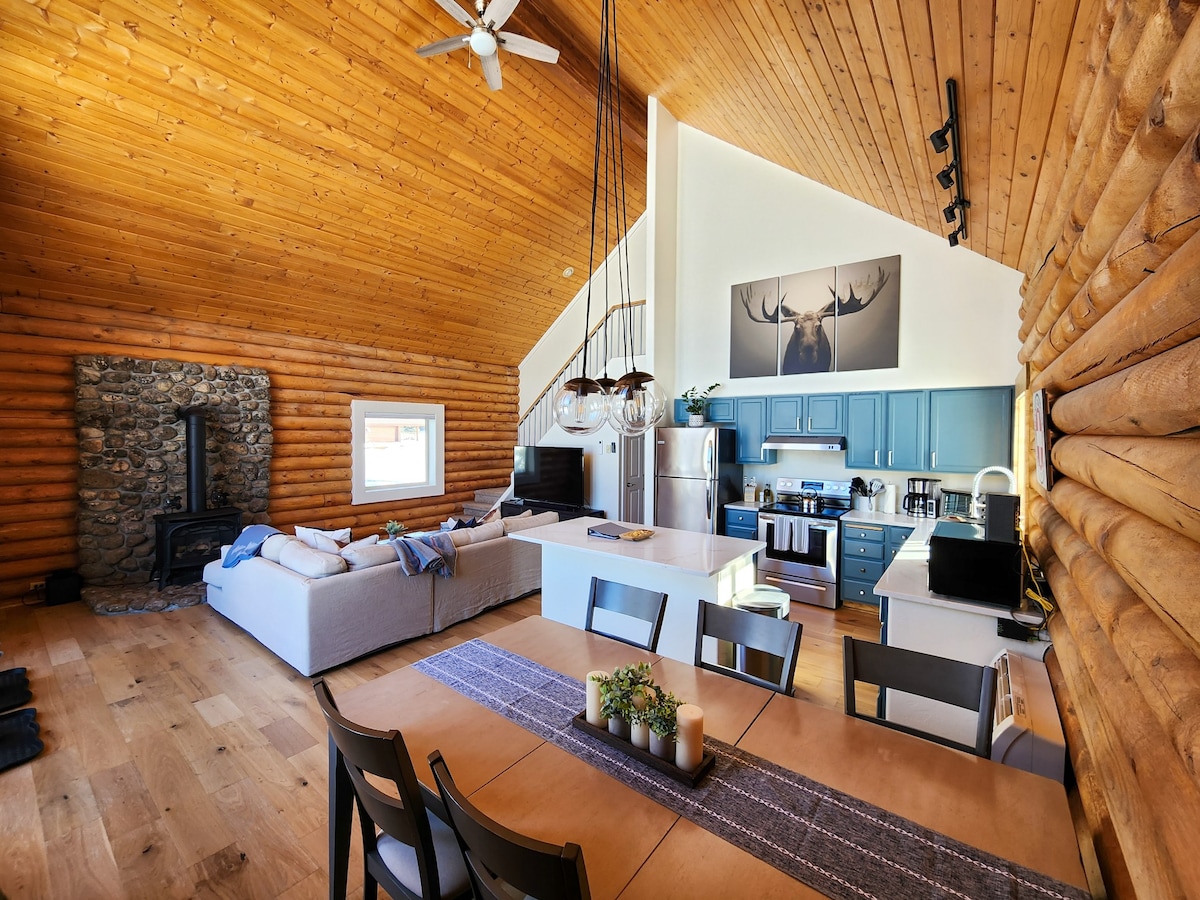 An open living space features high wooden ceilings and a cozy seating area with light-colored sofas. A dining table is set for six, complemented by a centerpiece of candles. The kitchen is equipped with modern appliances and a decorative moose art piece hangs above.