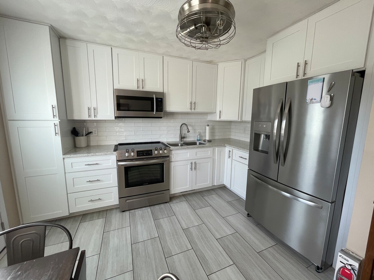 A modern kitchen features sleek white cabinetry and stainless steel appliances, including a large refrigerator and oven. A light-colored tile floor enhances the space, while a neutral backsplash complements the overall design. Ample counter space is available for meal preparation.