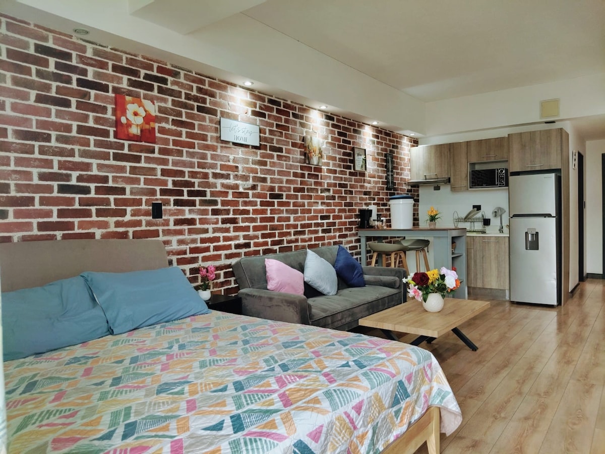 A modern living space features a cozy bed with a patterned blanket, softening the warm-toned wooden floor. A brick wall adds texture, complemented by ambient lighting. A comfortable sofa and coffee table create a welcoming area, while an equipped kitchenette is organized in the background.