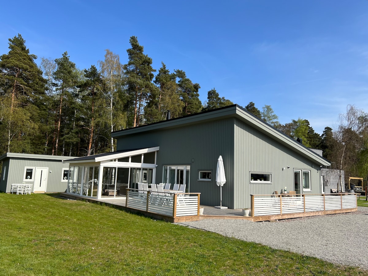 A modern villa is positioned within a green landscape, featuring a light grey exterior. A spacious deck extends from large glass doors, surrounded by trees. A gravel area is visible for parking, with outdoor seating options available nearby.
