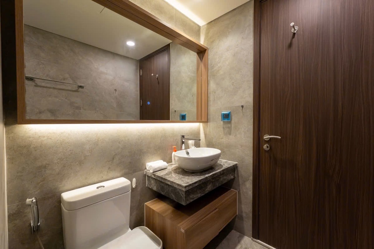 A modern bathroom is featured, showcasing a sleek countertop with a round sink. Warm lighting highlights the textured walls. A wooden cabinet provides storage beneath the sink, and a large mirror is mounted above. The toilet is positioned discreetly, contributing to the clean design.