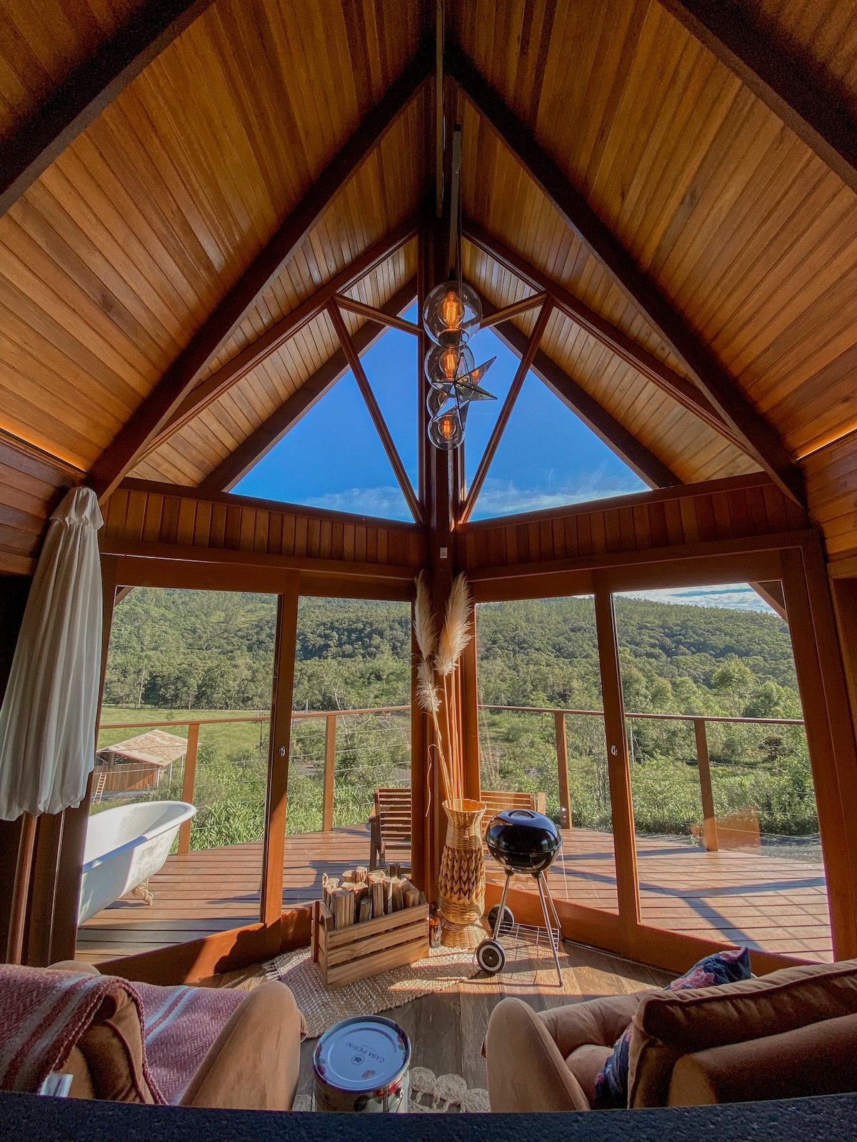 A cozy interior features large glass doors that frame a stunning view of the surrounding nature. A Victorian bathtub is positioned in front, with wooden beams accentuating the high ceiling. A compact grill and a decorative basket add functionality to the inviting living space.