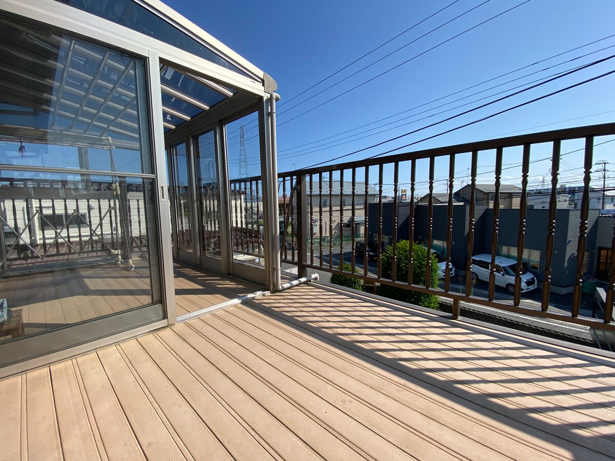 A spacious balcony features wooden flooring and a glass enclosure, allowing ample natural light. The surrounding area includes clear views of the neighborhood and parked vehicles. Railings provide safety while allowing unobstructed scenery.