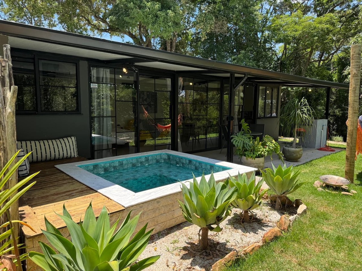 The exterior view of a modern chalet is presented, featuring a private, heated pool surrounded by landscaped greenery. Large glass doors open to an inviting interior space, visible in the background, enhancing the connection to nature.