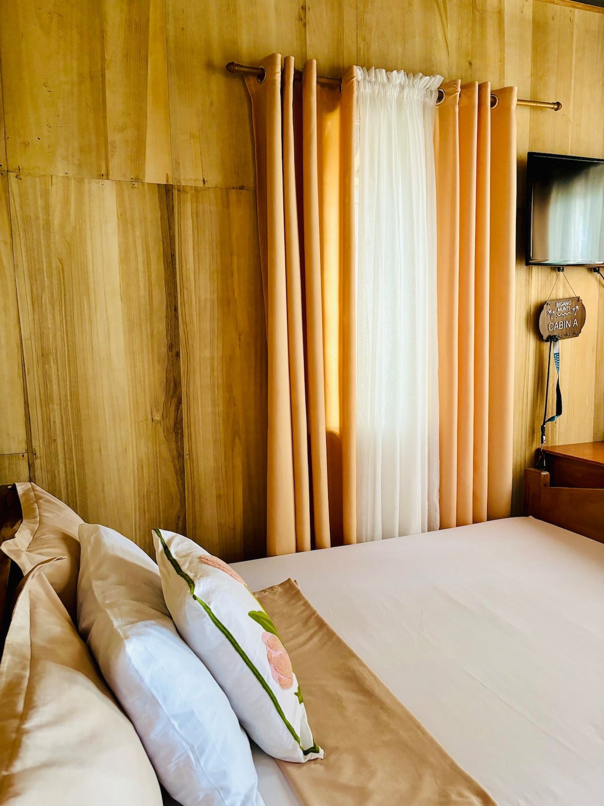 A cozy sleeping area features a spacious bed adorned with neutral-toned linens and decorative pillows. Soft curtains in warm hues frame the window, allowing natural light to brighten the room. A flat-screen TV is mounted on the wall above a wooden dresser.