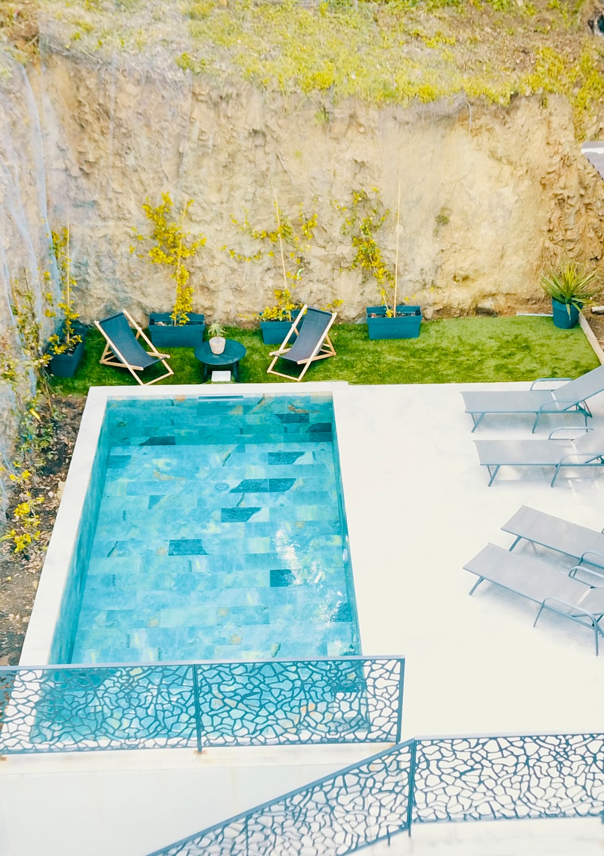 A modern pool area features a turquoise swimming pool surrounded by a stone deck. Lounge chairs are arranged around the pool, with several potted plants adding greenery. The backdrop consists of a natural stone wall dotted with yellow flowers, enhancing the serene outdoor environment.