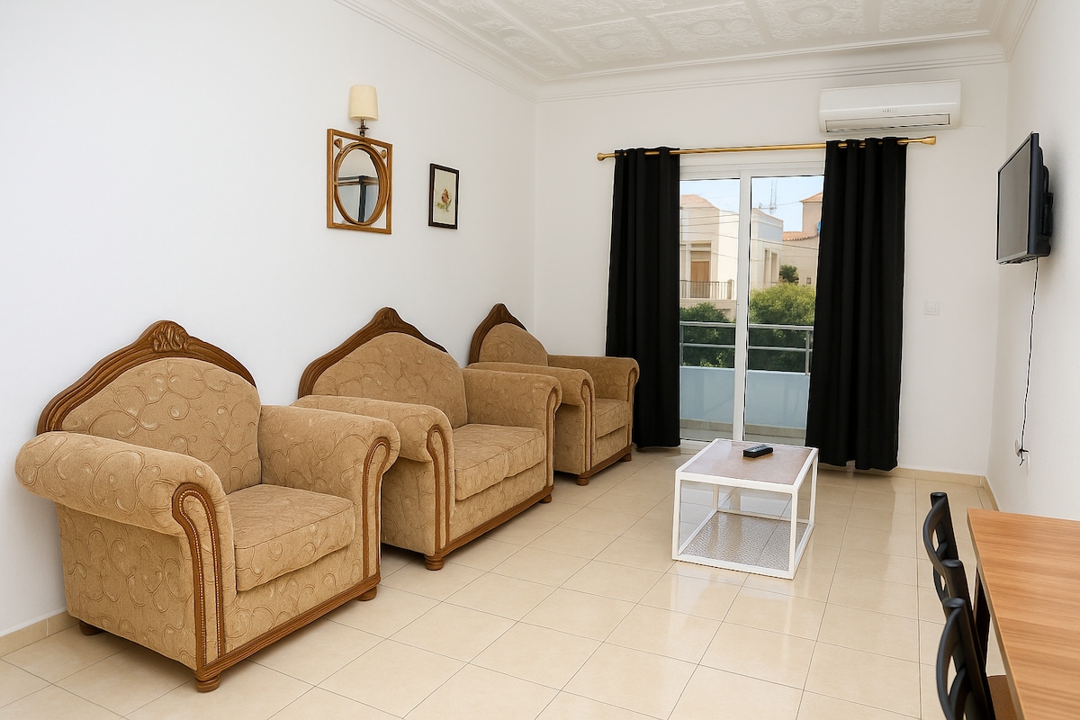 A bright and airy living area features two comfortable sofas arranged around a glass coffee table. Natural light enters through the glass door, leading to a balcony, while contrasting dark curtains frame the window. A television is mounted on the wall opposite the seating.