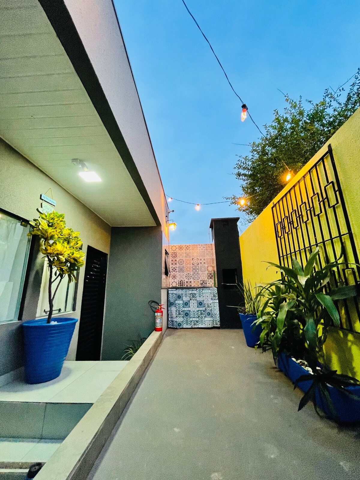 A well-lit outdoor pathway leads to a colorful wall adorned with intricate tiles. Green plants are positioned in blue pots, adding vibrancy. String lights hang overhead, gently illuminating the area, creating a welcoming ambiance for relaxation and social gatherings.