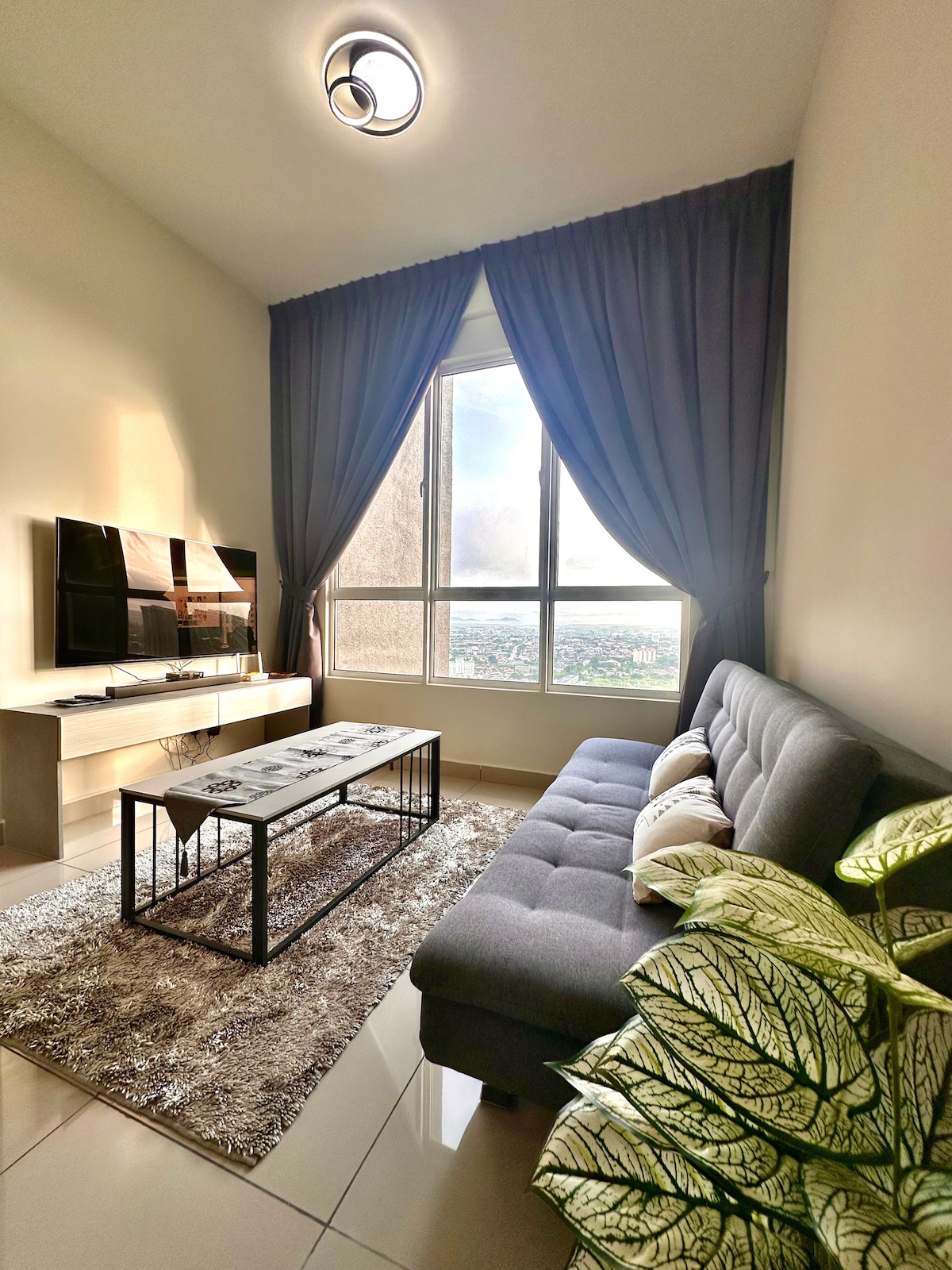 A comfortable sitting area is featured, highlighted by a sleek gray sofa and a coffee table positioned on a soft rug. A large window offers views of the surroundings, with natural light filling the space. Decorative plants add a touch of greenery.