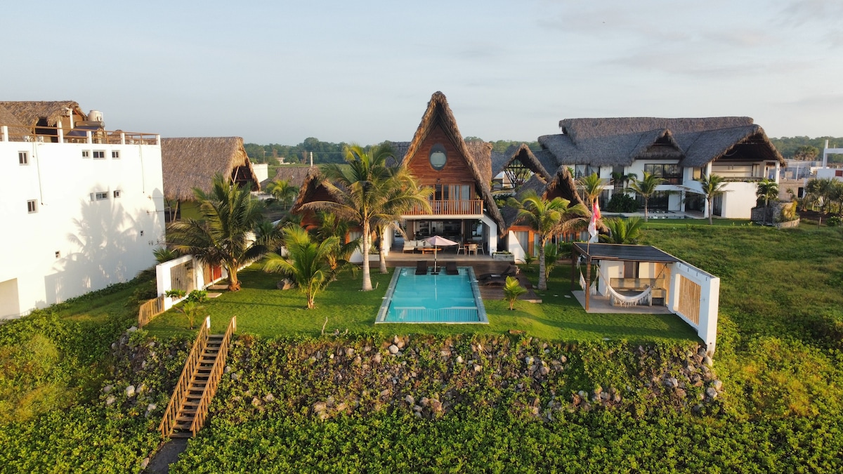 An aerial view captures a spacious property featuring a main house with a distinct architecture and a thatched roof. A crystal-clear pool is centered within a lush green landscape. Surrounded by palm trees, the outdoor space invites relaxation and enjoyment of the natural environment.