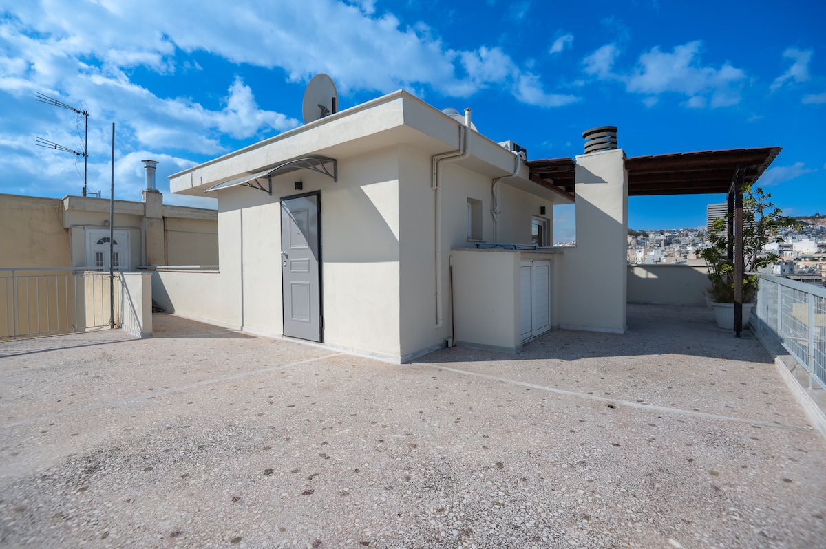 A spacious rooftop area is featured with a smooth, light-colored surface and a satellite dish visible on the building's edge. The clear blue sky enhances the setting, while parts of the surrounding urban landscape can be glimpsed in the background.