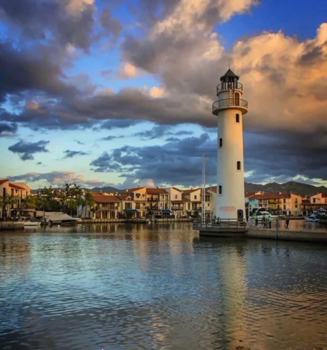 A lighthouse stands by the water's edge, surrounded by pastel-colored buildings. The calm water reflects the vibrant sky filled with clouds, creating a serene atmosphere. Boats are visible in the distance, suggesting a lively maritime environment.
