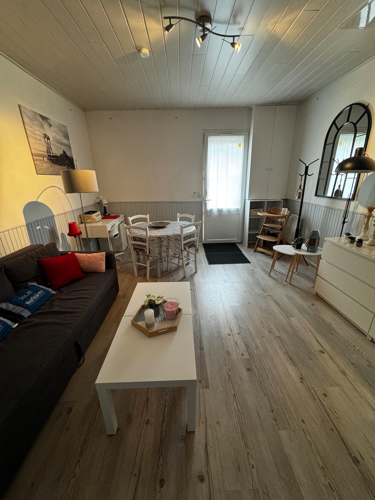 A well-lit living area features a dark sofa adorned with decorative pillows and a light coffee table. A dining table with four chairs is set against a wall. Natural light streams through a door with a sheer curtain, illuminating the wooden flooring.