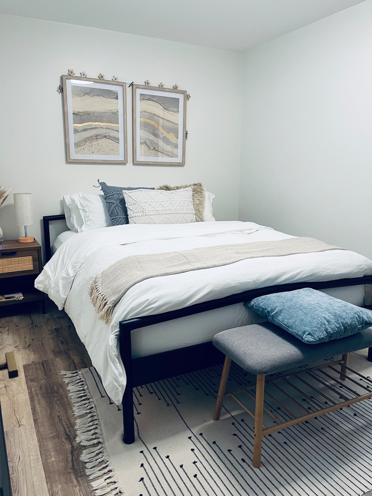 A neatly arranged bedroom features a cozy bed adorned with soft pillows and blankets. Framed artwork hangs above, complemented by a bedside lamp and woven storage. A small stool is positioned near the bed, and a plush area rug covers the floor, enhancing the space’s inviting ambiance.