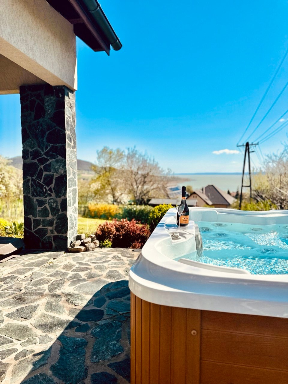 An outdoor jacuzzi is surrounded by a stone patio, offering a view of the lush landscape and distant hills. A bottle of wine is placed on the edge of the tub, while bright blue skies and sunlight create a serene atmosphere.