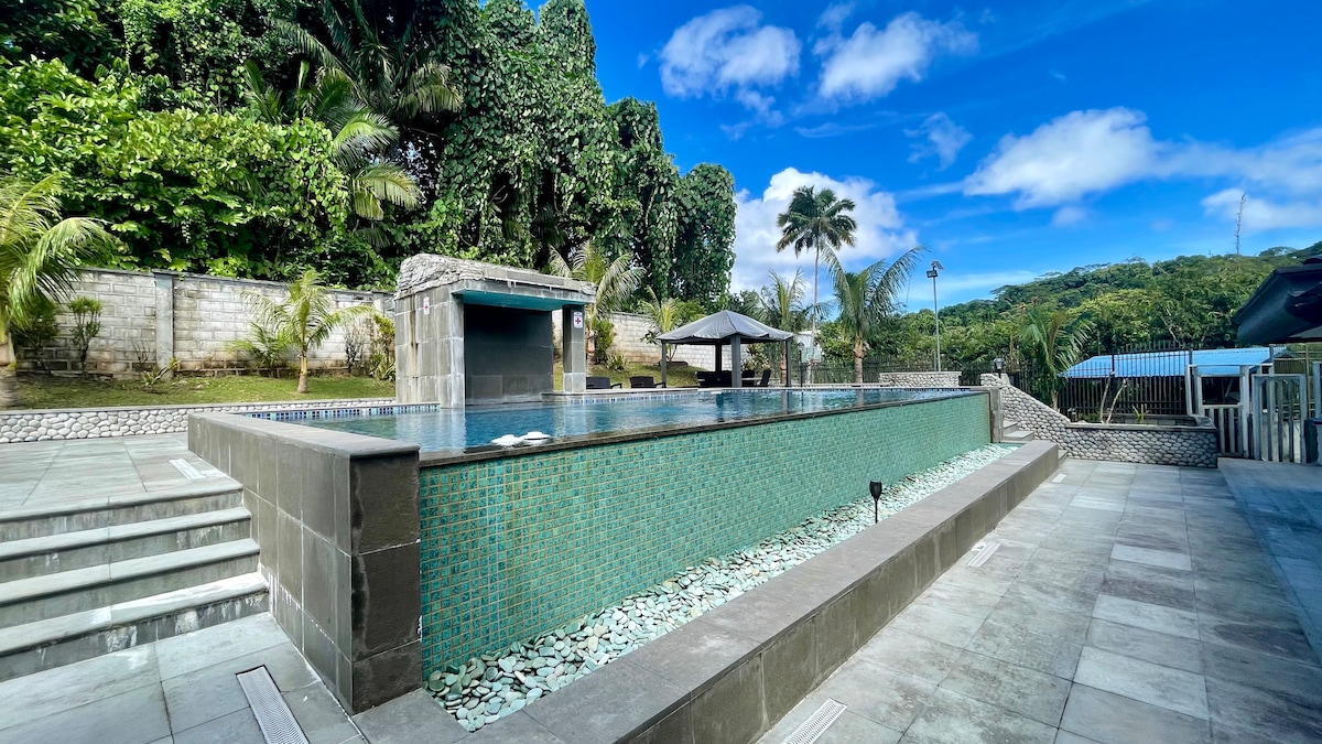An infinity pool is framed by stone tiles and surrounded by lush greenery. The tranquil water reflects the clear blue sky, while shaded seating areas are available on one side, creating a serene outdoor space for relaxation.