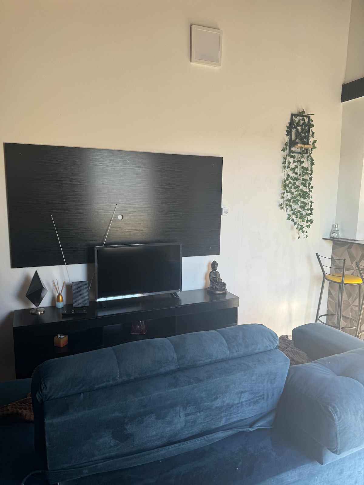 A seating area features a dark blue sofa facing a minimalist black entertainment unit with a flat-screen television. Decorative elements include a small figurine and a plant accent on the wall, enhancing the modern aesthetic of the space.