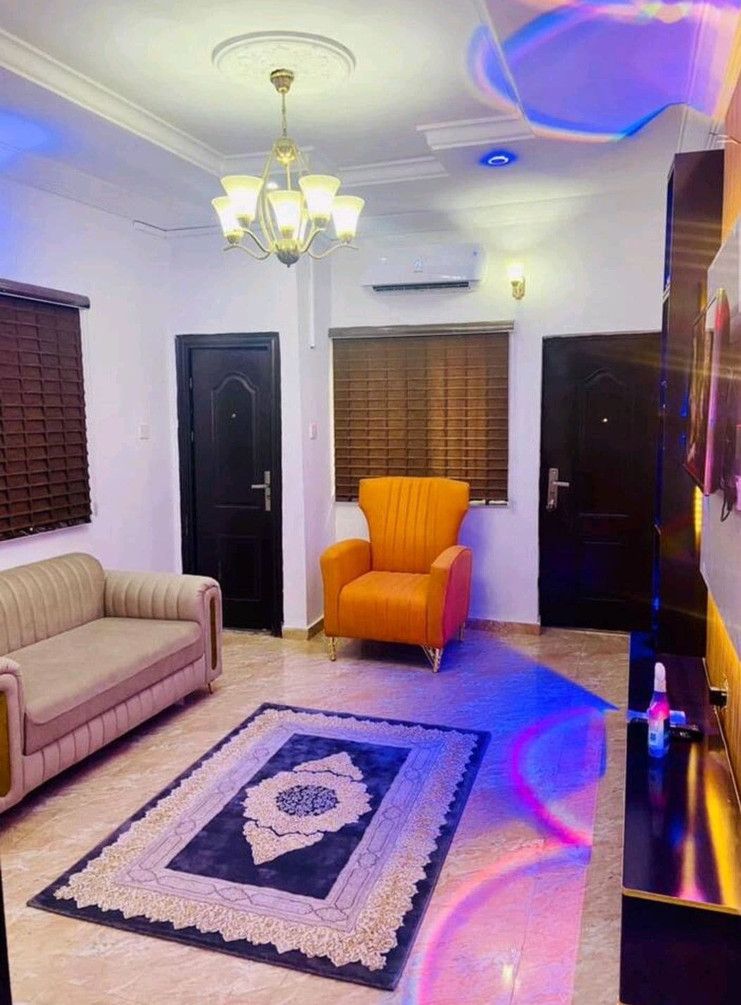 A stylish living area features a beige sofa and an orange armchair, creating a comfortable seating arrangement. The space is illuminated by a modern chandelier, while wooden blinds cover the windows, allowing for adjustable light levels. A patterned rug adds warmth to the room.
