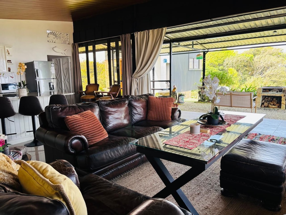 A spacious living area features dark leather seating arranged around a modern glass table. Natural light floods the space through large sliding doors, opening to the outdoors. A small kitchenette is visible in the background, accompanied by light neutral decor and decorative elements.