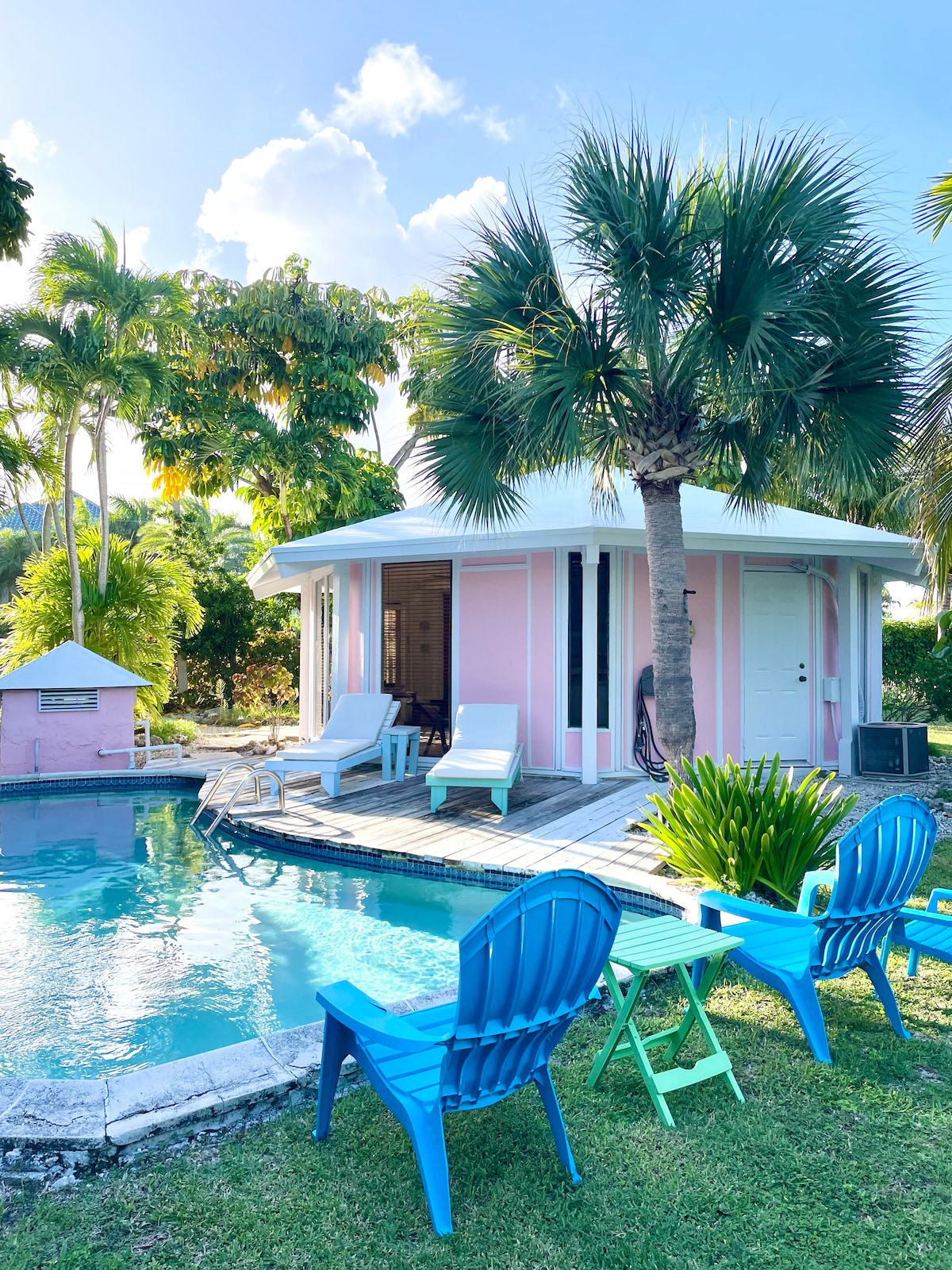 A charming cottage with a circular design is surrounded by lush greenery and palm trees. A serene swimming pool sits in the foreground, flanked by colorful adirondack chairs. The cottage features light pink walls and large windows, inviting natural light into the interior.