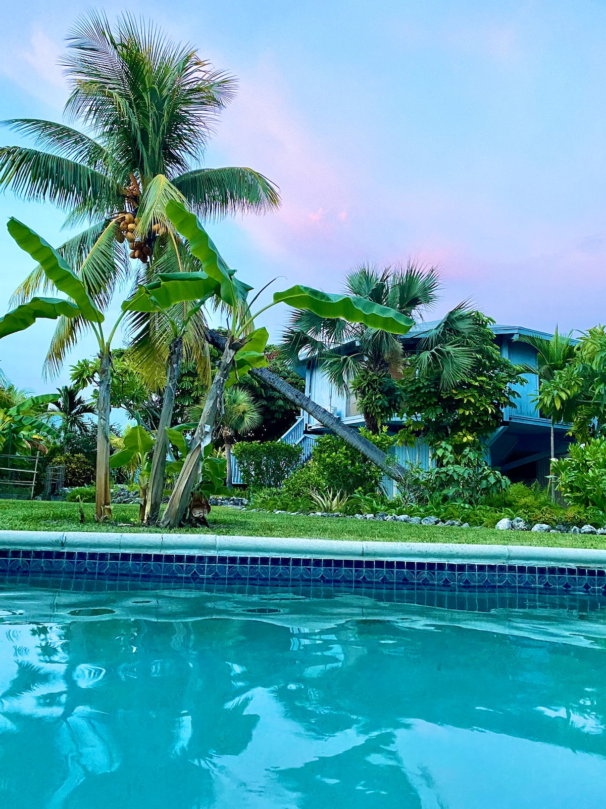 The image captures a serene view of the pool with clear water, reflecting the sky's soft colors. Lush tropical plants, including palm and banana trees, surround the pool, while a welcoming cottage is seen in the background, nestled among vibrant greenery.