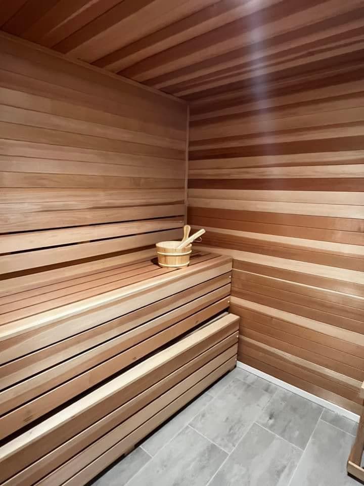 A wooden sauna interior is presented, featuring smooth cedar walls with horizontal slats. Wooden benches are arranged for seating, and a small wooden bucket with a ladle sits atop a bench, providing a serene environment.