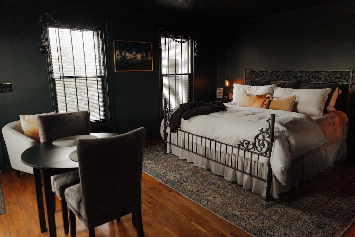 The studio features a king-sized bed with an elegant metal frame, complemented by soft linens and decorative pillows. A cozy seating area with a round table and two chairs sits near the window, which is framed by curtains. Warm lighting enhances the intimate ambiance.
