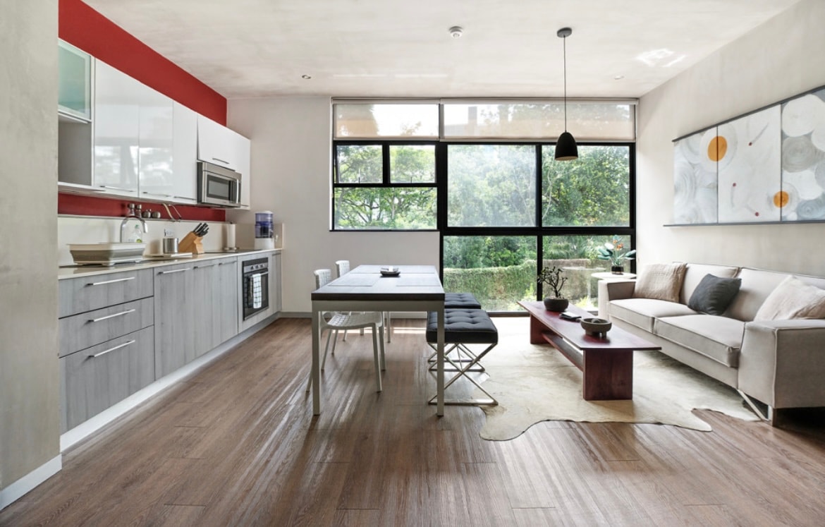 A bright, open living area features large windows allowing natural light to flood the space. The kitchen is equipped with modern appliances, complemented by a dining table and comfortable seating. A piece of artwork adorns the wall, while a soft rug adds texture to the wooden flooring.