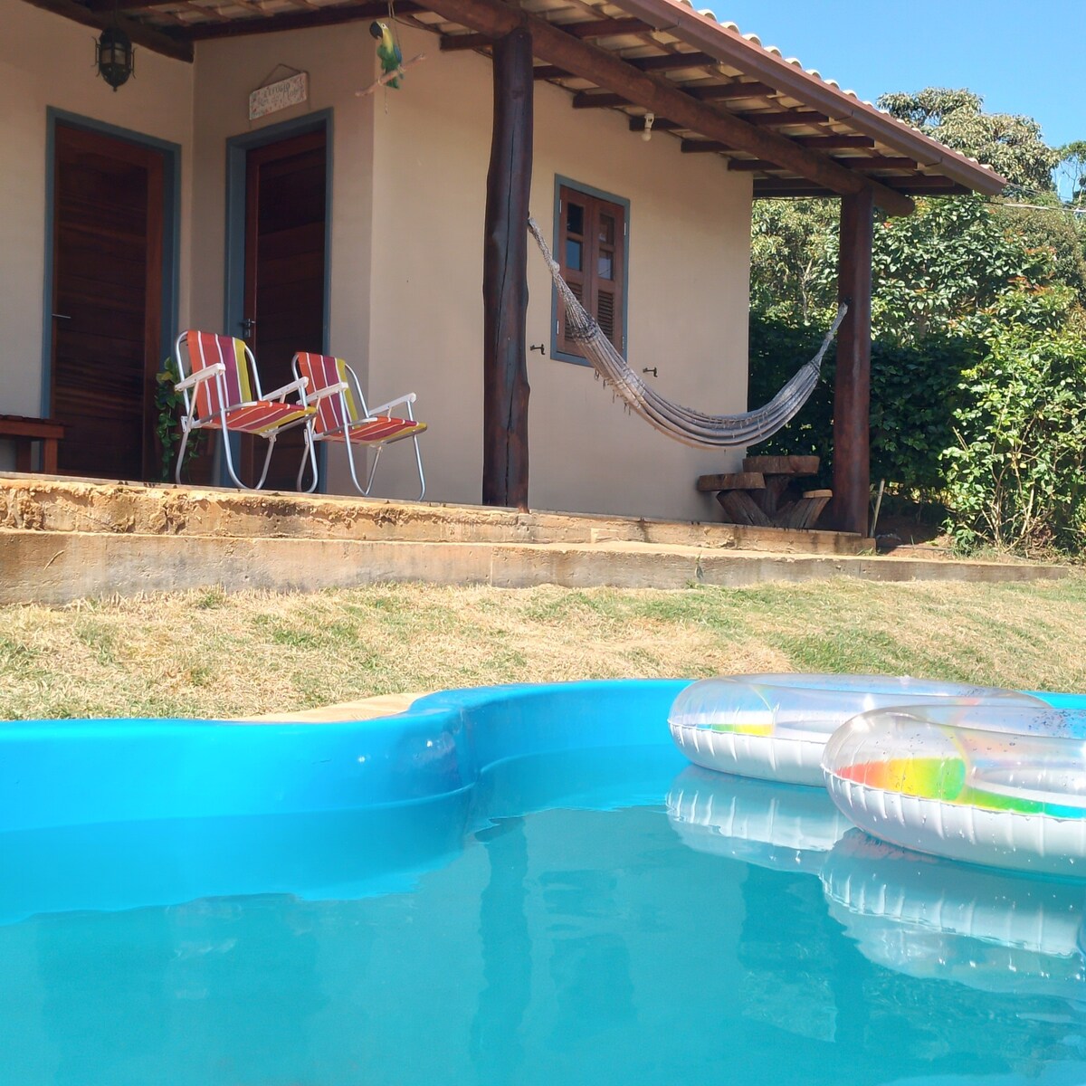 A private outdoor space features a small pool with colorful floats resting on the surface. Two striped chairs sit on a porch under a wooden roof, alongside a hammock swaying gently. The lush greenery surrounds the area, enhancing the natural setting.