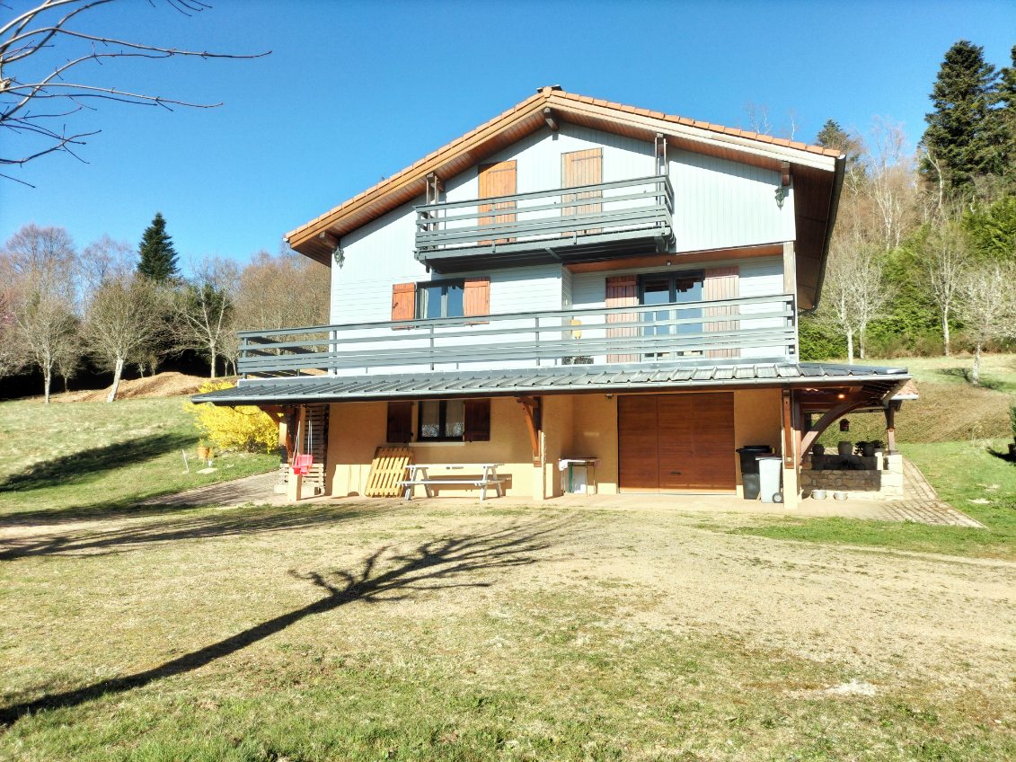 A spacious chalet is depicted, featuring a light-colored exterior with multiple balconies. The well-maintained grounds showcase a grassy area surrounded by trees, providing a serene environment. A covered patio area below offers seating options, and a large driveway is visible, accommodating multiple vehicles.