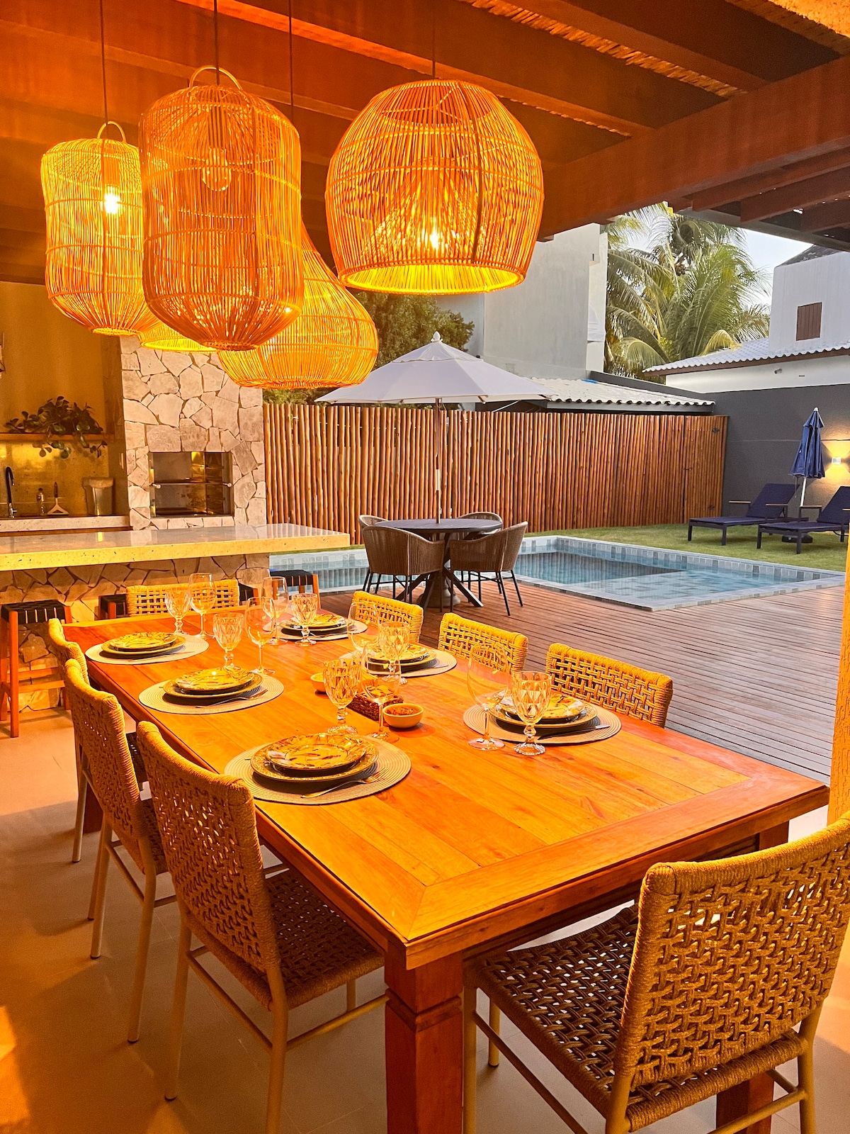 An inviting dining area is highlighted by woven pendant lights overhead. A wooden table, set for dinner, features plates, glasses, and utensils. A cozy ambiance is complemented by a view of a pool and outdoor seating in the background, surrounded by natural greenery.