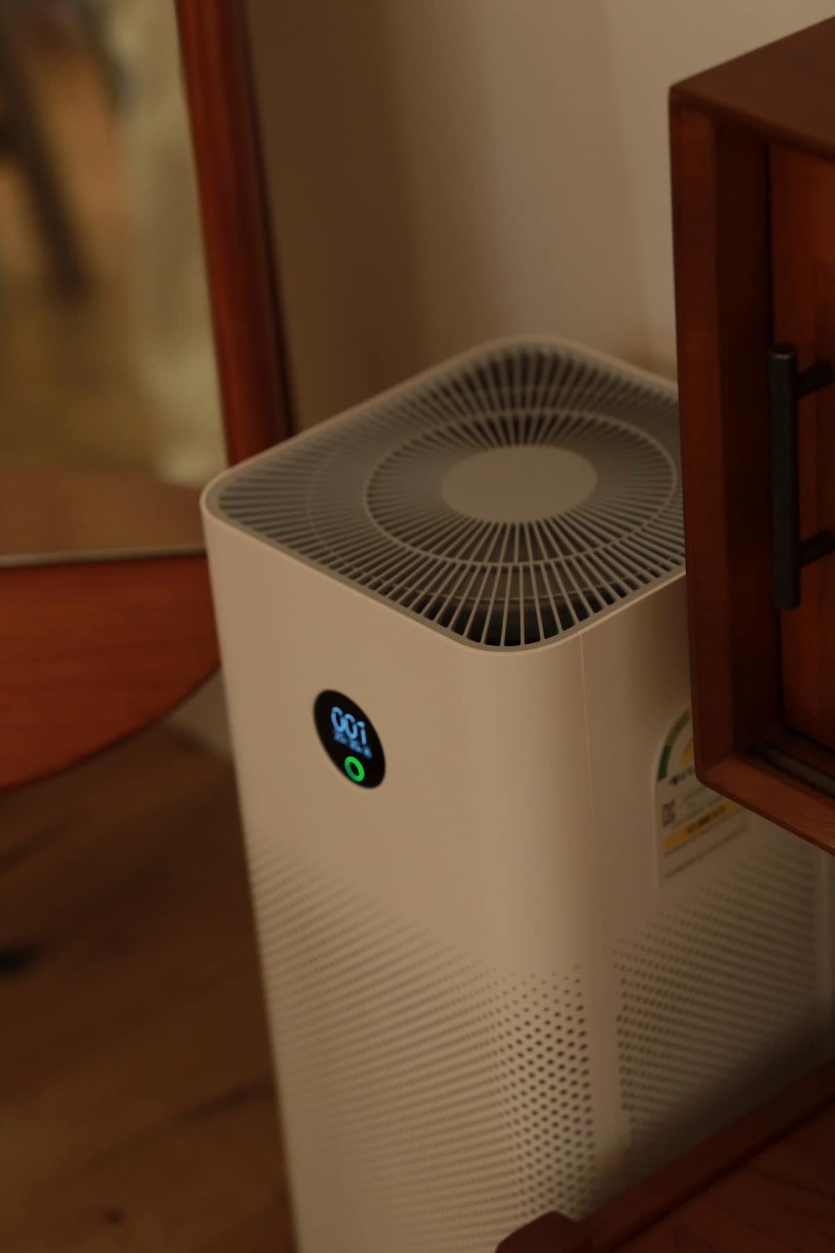An air purifier is positioned between a wooden cabinet and a mirror, featuring a circular display that indicates the current air quality. The device has a clean white design, contributing to a modern and functional aesthetic in the space.