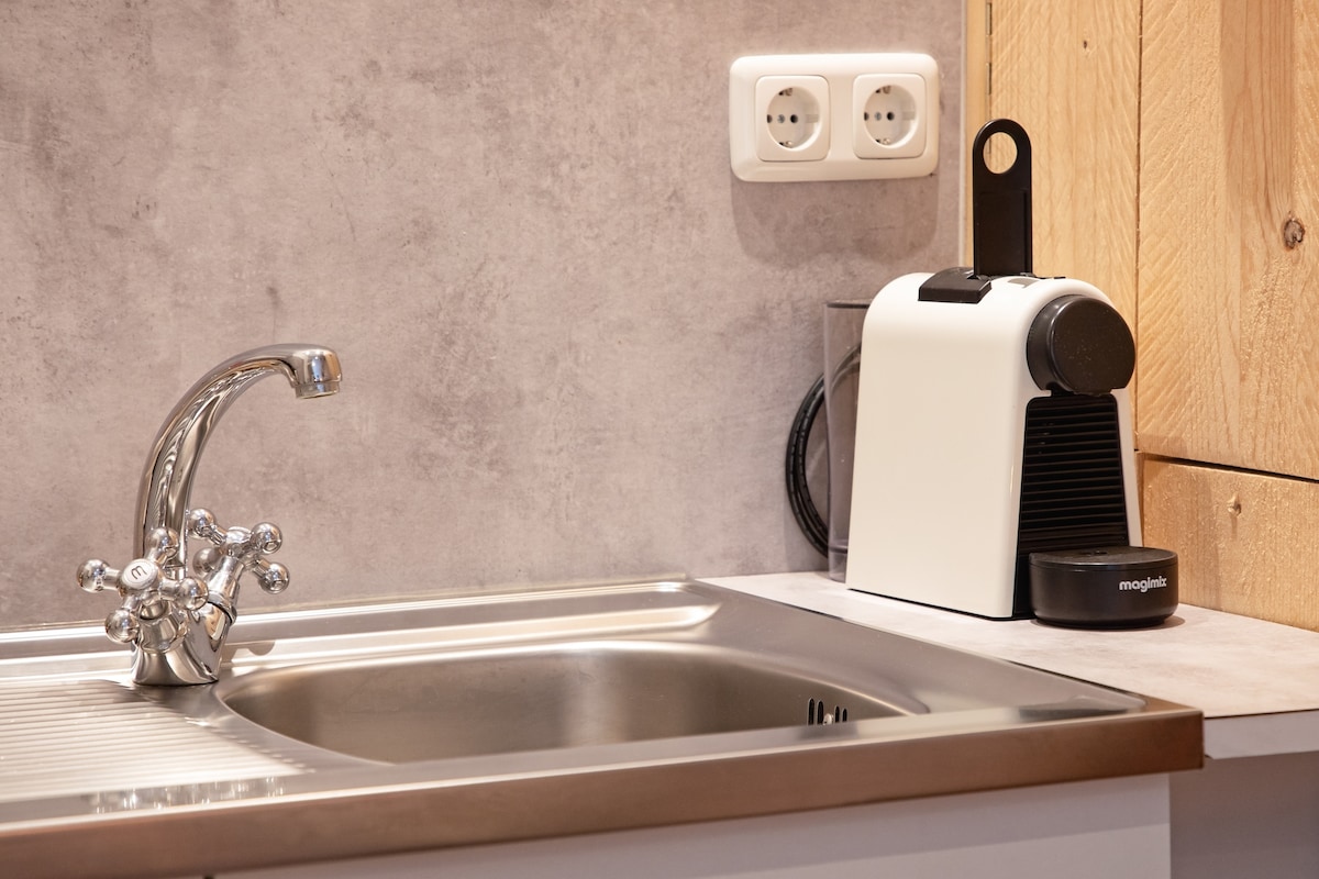 A modern kitchenette is highlighted, featuring a sleek metal sink with a stylish faucet. An espresso machine is positioned beside the sink, complemented by an electric outlet on the wall. The textured gray countertop adds a contemporary touch.