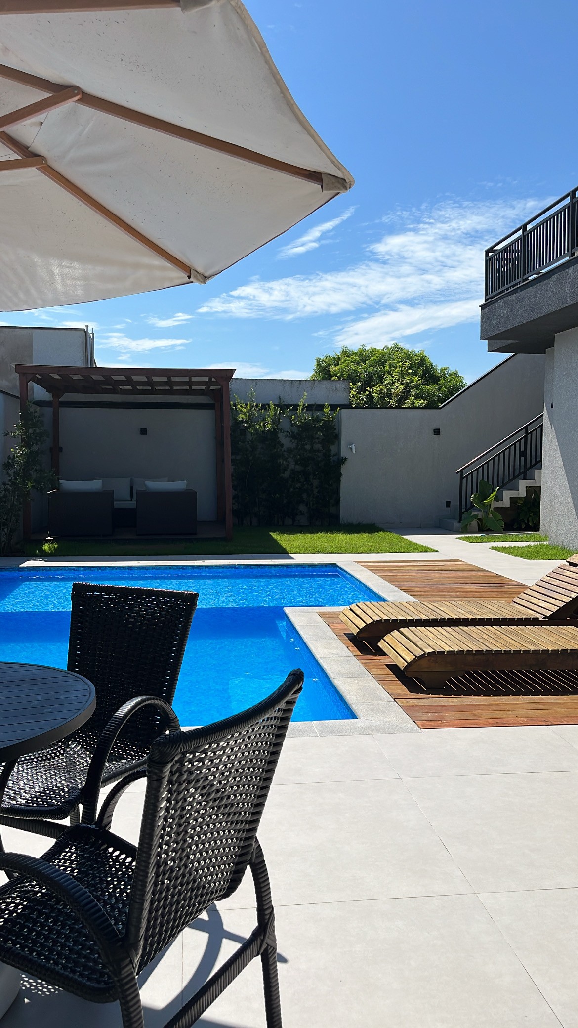 An outdoor space features a blue swimming pool surrounded by lounge chairs and a shaded seating area. A patio table with black woven chairs is positioned nearby. Lush greenery is seen in the background, along with a staircase leading to a second level.