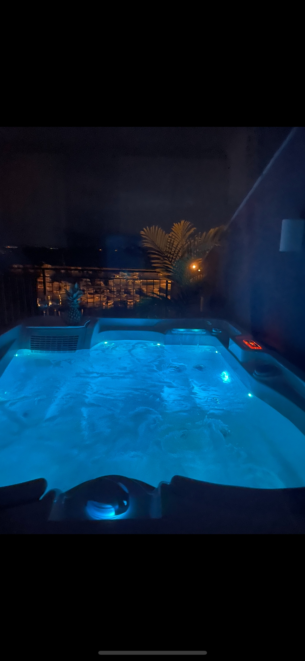 A hot tub is illuminated with soft blue lighting, inviting relaxation. The surrounding area is dimly lit, with silhouettes of plants and distant lights visible through a railing. The soothing water and tranquil ambiance create a serene space for guests.
