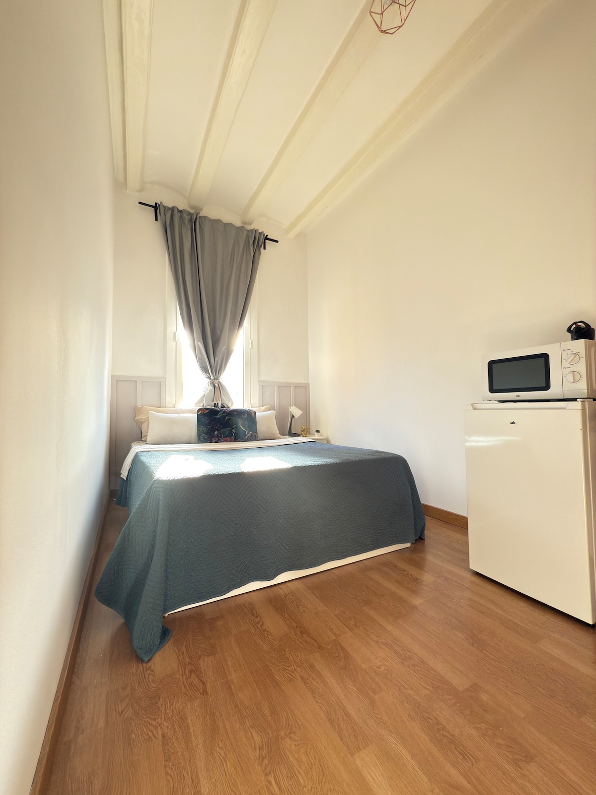 A well-lit sleeping area is featured, showcasing a spacious bed covered with a textured blue blanket. A small white fridge and microwave are positioned near the wall, while natural light enters through the window, framed by elegant gray curtains.
