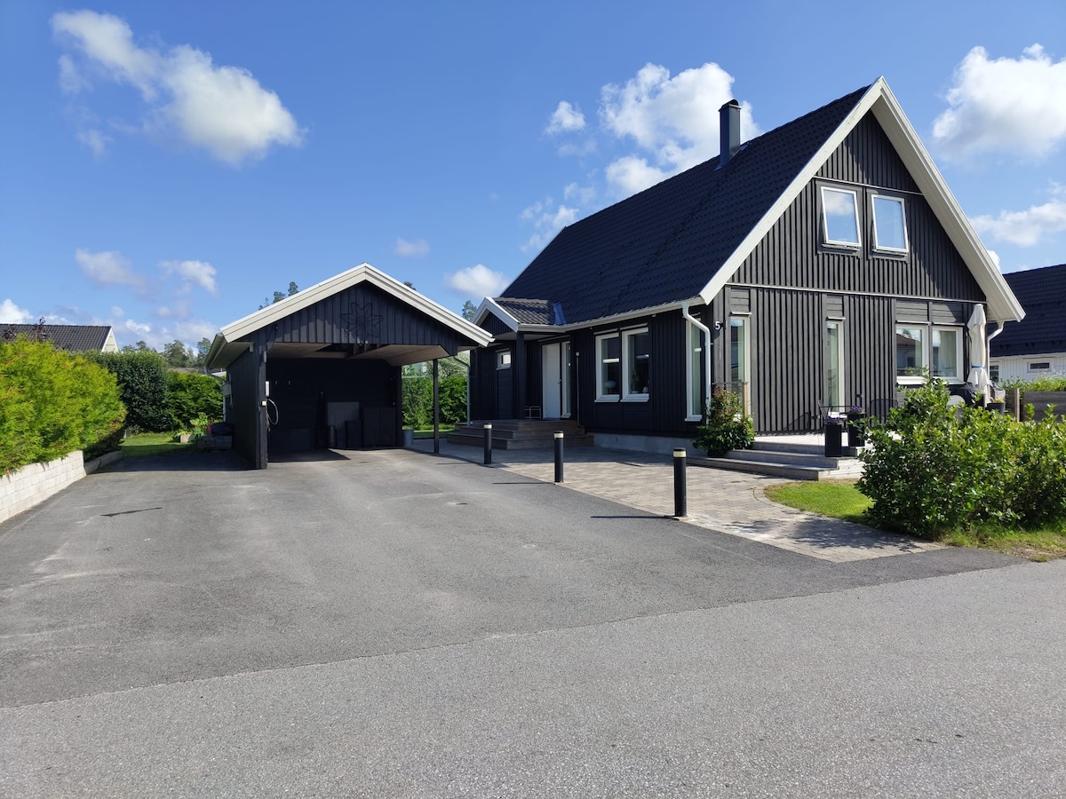 The image showcases a modern two-story house with black wooden siding and a sloped roof. A spacious driveway leads to a covered carport on the left, surrounded by greenery. The clear sky above highlights the calm residential area.