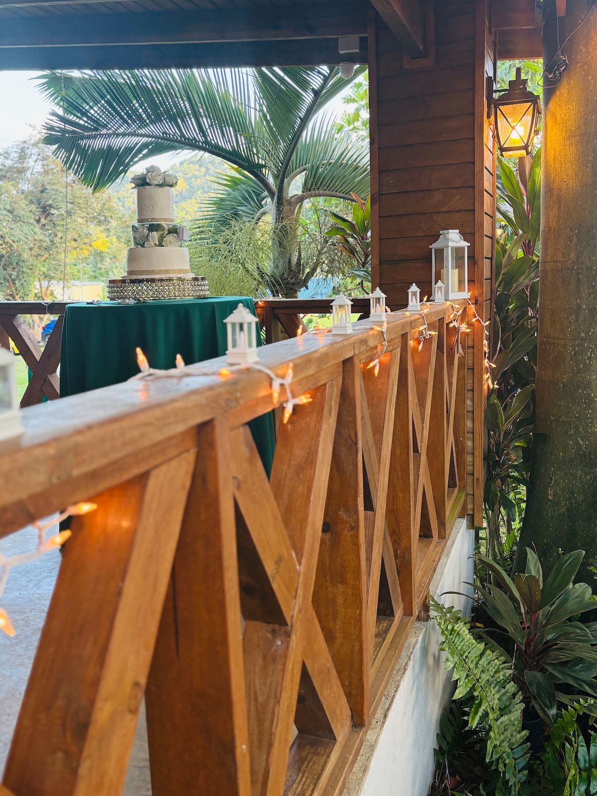 A wooden railing lined with warm string lights creates a welcoming ambiance. A table draped in green cloth is visible in the background, featuring a decorative cake. Lush greenery and palm leaves frame the scene, enhancing the natural setting.
