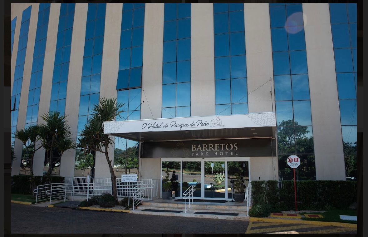 The hotel's exterior features a modern facade with large glass windows that reflect the surrounding greenery. An accessible entrance is highlighted by a ramp, and the hotel name 'Barretos Park Hotel' is prominently displayed above the entrance.